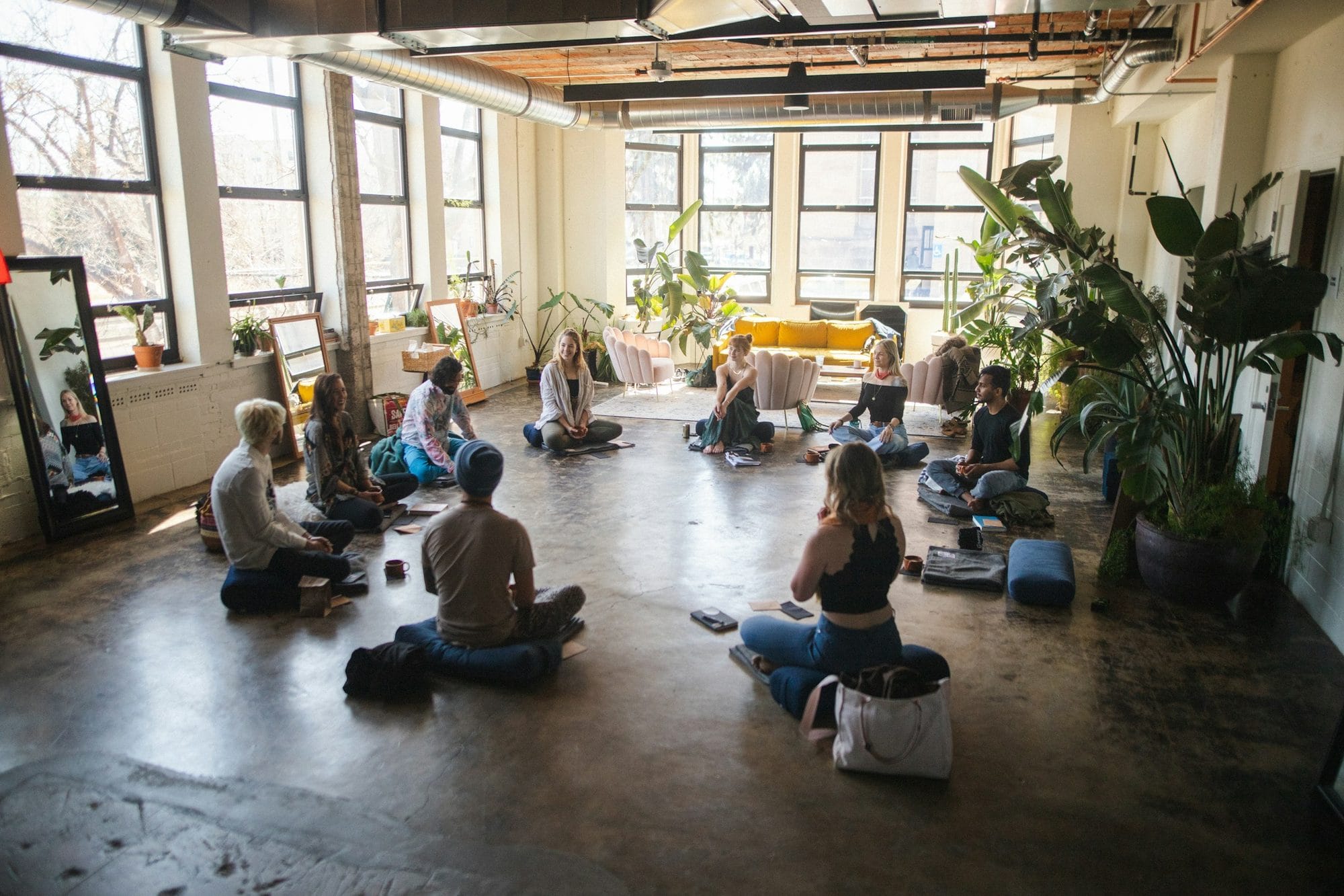 A small group sitting in a circle on cushions in a bright, plant-filled studio
